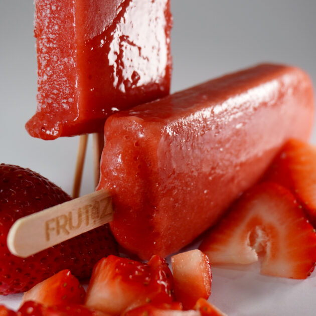 Paletas de fresa artesanales sobre un fondo blanco, acompañadas de fresas frescas y trozos de fresa, destacando su color rojo brillante y textura suave.
