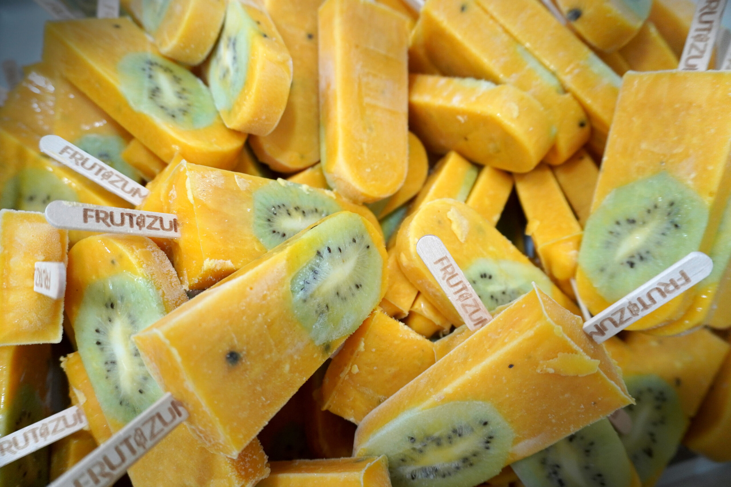 Paletas de frutas congeladas de color amarillo con rodajas de kiwi, presentadas en una caja. Las paletas llevan el nombre "FRUTIZUN" en los palitos de madera. Ideal para refrescarse en verano.